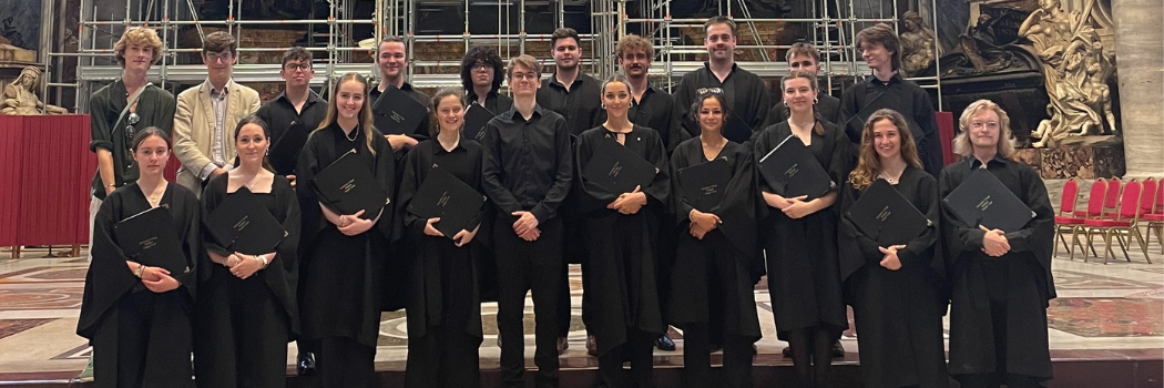 Group of students gowned in black robes standing in a choir formation holding music folders. They are standing in St Peter’s Basilica.