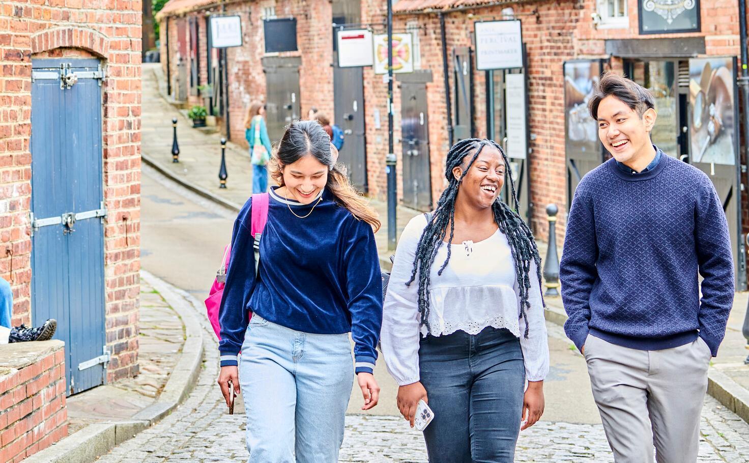 Students walk along a cobbled road - they are chatting and are dressed in jumpers and long sleeved tops and it is quite overcast. Behind them are red brick buildings with black wooden shutters. Taken in Back Silver Street, also known as Fowler's Yard, in the City centre.