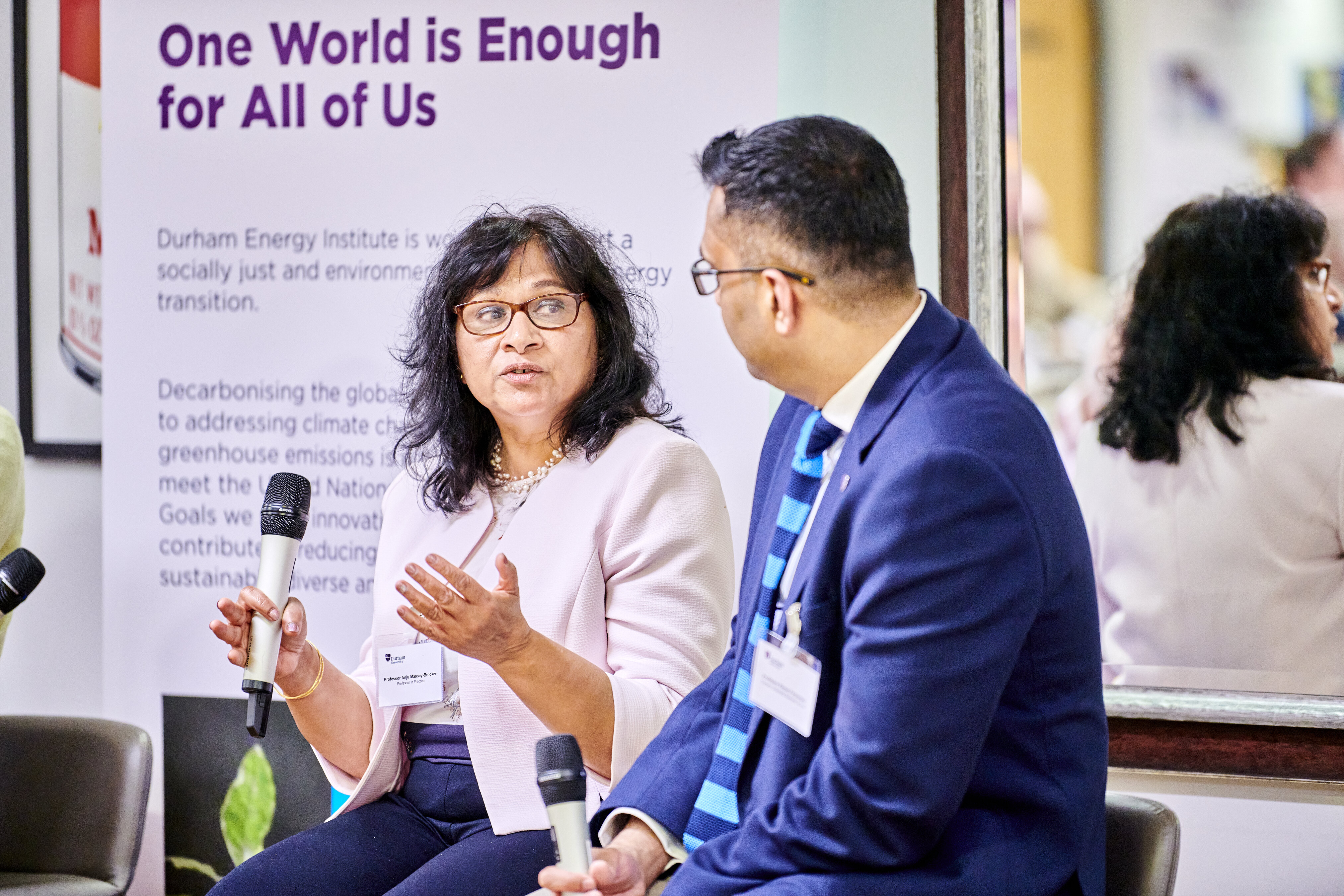 A smartly dressed woman with a microphone talking and making hand gestures looking at a man in a suit sitting next to her