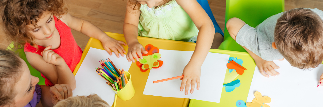 Young children sitting at a classroom table with the teacher doing crafts
