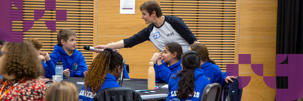 A woman in a black and white top holds a microphone towards a school boy in a blue jumper.