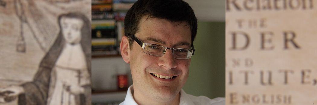 Head and shoulders close-up of a man smiling directly at the camera. He has short dark hair and is wearing glasses.