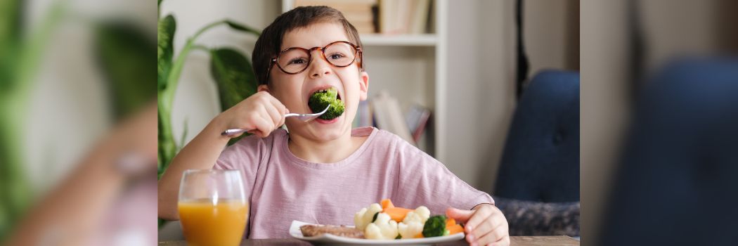 A young boy wearing glasses and sitting at a table eats a green vegetable from a plate full of vegetables.
