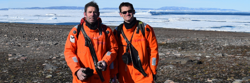 Professors Colm O’Cofaigh and Dave Roberts, wearing orange jumpsuits, standing on an artic beach