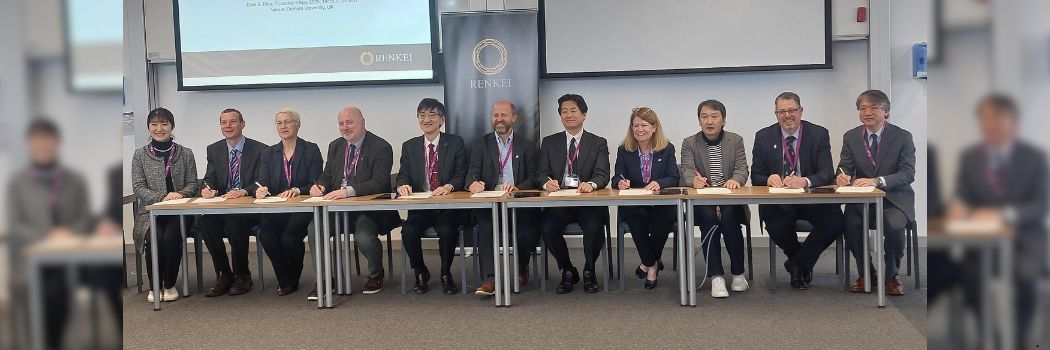 A row of people sitting behind tables and signing documents. A banner behind them reads RENKEI.