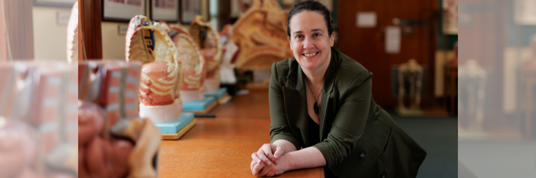 Professor Siân Halcrow smiling wearing a dark green blazer and leaning on a table in a seminar room