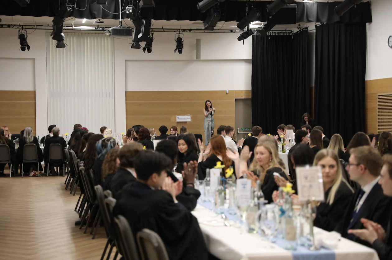 Students seated at long formal tables applaud a speaker standing at a microphone during a college awards dinner at John Snow College. The room is warmly lit, with daffodils and table settings creating a celebratory atmosphere.