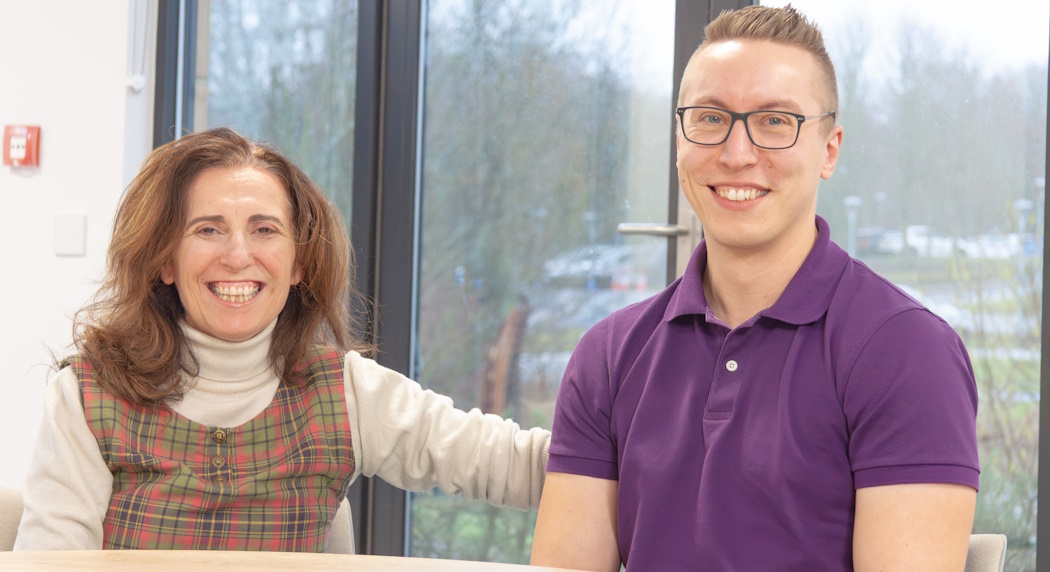A photo of two people sat at a table in a meeting room looking at the camera.