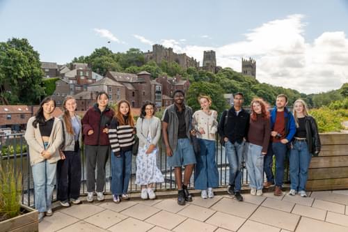 A large group of students by the riverside with the castle and cathedral in the background