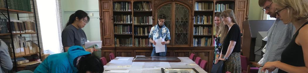 A group of students are standing around a table in the centre of the room, which is covered in bubble wrap. On top of this are framed images of artworks, which the group is looking at. One of the group has leaned over the table for a closer look.